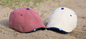 The Declaration Scally Cap is here in Faded Red and Vintage White, both with a blue and white star lining, shown sitting on a beach